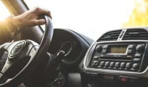 Close-up of a hand on a steering wheel inside a car, dashboard visible.